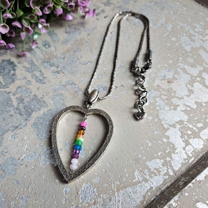 May include: A silver heart-shaped pendant necklace with a rainbow of coloured beads hanging inside. The necklace has a silver chain.