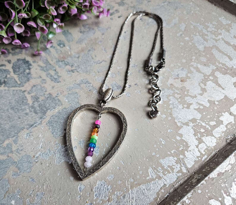May include: A silver heart-shaped pendant necklace with a rainbow of coloured beads hanging inside. The necklace has a silver chain.