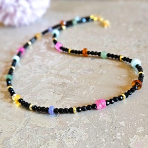 May include: A beaded necklace featuring black, gold, and colorful beads. The necklace has a black cord with small black beads, interspersed with gold accents and vibrant beads in pink, orange, green, and blue.