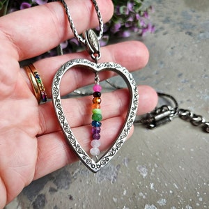 May include: A silver heart-shaped pendant necklace with a chain. The pendant has a detailed design and features a dangling row of colourful gemstones.