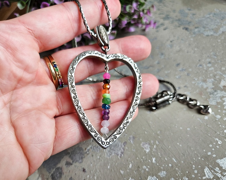 May include: A silver heart-shaped pendant necklace with a chain. The pendant has a detailed design and features a dangling row of colourful gemstones.