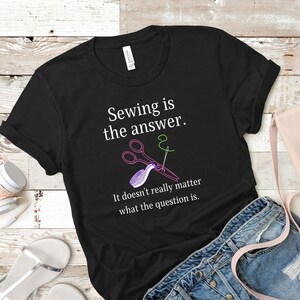 以下が含まれることがあります： 黒いTシャツに白い文字で「縫うことは答え。どんな質問でも関係ない。」と書かれています。Tシャツには、ピンクのハサミ、針と糸、メジャーが描かれています。