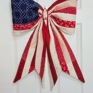 May include: A large fabric bow featuring a patchwork design with red, white, and blue colors. The left side displays a blue field with white stars, while the right side and tails feature red and white stripes with various patterns. Text is visible.