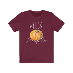 May include: A burgundy t-shirt featuring the text "HELLO Pumpkin" in white script, with a watercolour-style orange pumpkin beneath. The shirt has a crew neck and short sleeves.