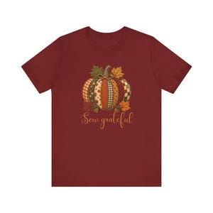 Może przedstawiać: Ciemnoczerwony t-shirt z krótkim rękawem z centralną grafiką dyni wykonaną z różnych wzorzystych segmentów tkaniny. Dynia jest ozdobiona jesiennymi liśćmi i żołędziami, a poniżej znajduje się złoty napis Sew grateful.