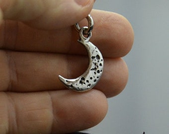 May include: A silver crescent moon charm with a textured surface. The charm is a simple design with a smooth, rounded edge.
