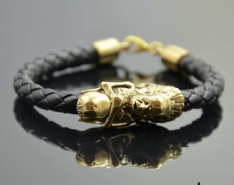May include: Black braided leather bracelet with a gold-colored decorative clasp featuring a detailed sculpted design. The bracelet has gold-colored end caps and a matching clasp.