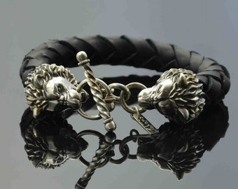 May include: A black leather bracelet with silver lion head clasps. The bracelet is braided and has a silver toggle clasp.