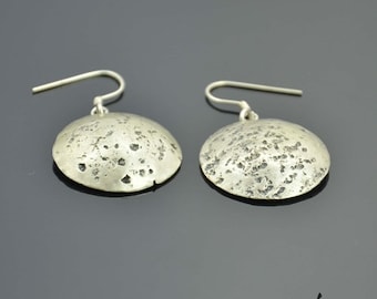 May include: A pair of silver-toned earrings. Each earring features a textured, disc-shaped pendant with a hammered appearance, suspended from a simple hook. The earrings are displayed against a dark gray background.