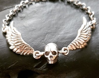 May include: A silver bracelet with a skull charm and two wings on either side. The bracelet is made of a chain link design.