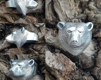 May include: A silver ring with a bear head design. The bear's head is detailed with fur and has a realistic expression. The ring is shown on a wooden background.
