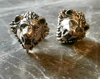 May include: A pair of silver cufflinks featuring detailed lion head sculptures. The lion heads are facing forward with their mouths open, showing their teeth.