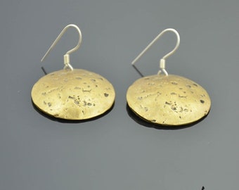 May include: A pair of gold-colored, disc-shaped earrings with silver-colored ear wires. The earrings have a textured surface and a hammered appearance. The earrings are displayed on a dark gray surface.