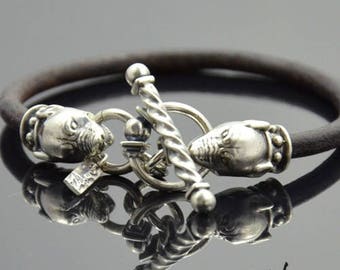 May include: A brown leather bracelet with a silver toggle clasp featuring two dog head designs. The clasp is twisted and has a small bead accent.