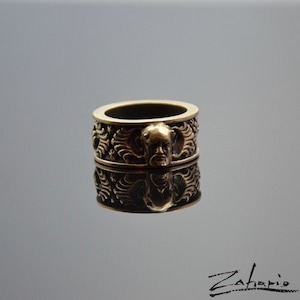 Puede incluir: Un anillo de color bronce con un diseño intrincado. El anillo presenta un relieve detallado de una cara con barba y alas estilizadas. El anillo mide aproximadamente 2,5 cm de ancho y tiene una superficie reflectante.