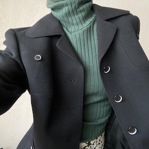 Puede incluir: Un abrigo negro con solapas y botones plateados. El abrigo se lleva sobre un jersey de cuello alto verde oscuro acanalado. El abrigo tiene un diseño clásico con un corte entallado, adecuado para diversas ocasiones.