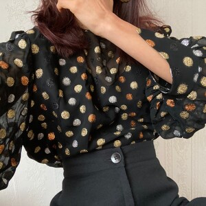 May include: A black sheer blouse with gold and silver polka dots. The blouse has long sleeves with ruched cuffs and a loose fit.