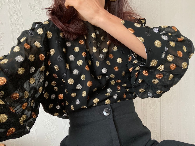 May include: A black sheer blouse with gold and silver polka dots. The blouse has long sleeves with ruched cuffs and a loose fit.