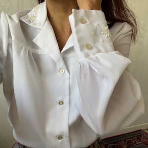 Puede incluir: Una blusa blanca de manga larga con cuello y puños con volantes. El cuello y los puños están decorados con adornos de perlas.