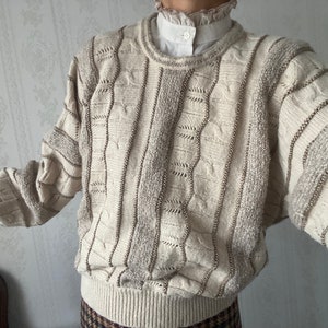 Puede incluir: Un suéter de punto color crema con un patrón texturizado y detalles de cuentas. El suéter tiene un cuello redondo y mangas largas.