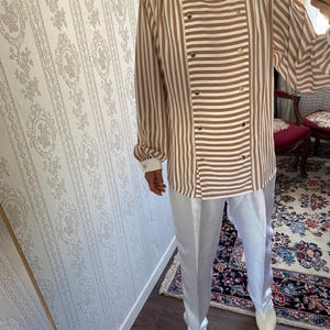May include: A brown and white striped long-sleeve shirt with a double-breasted button closure. The shirt has a relaxed fit and is paired with white pants.