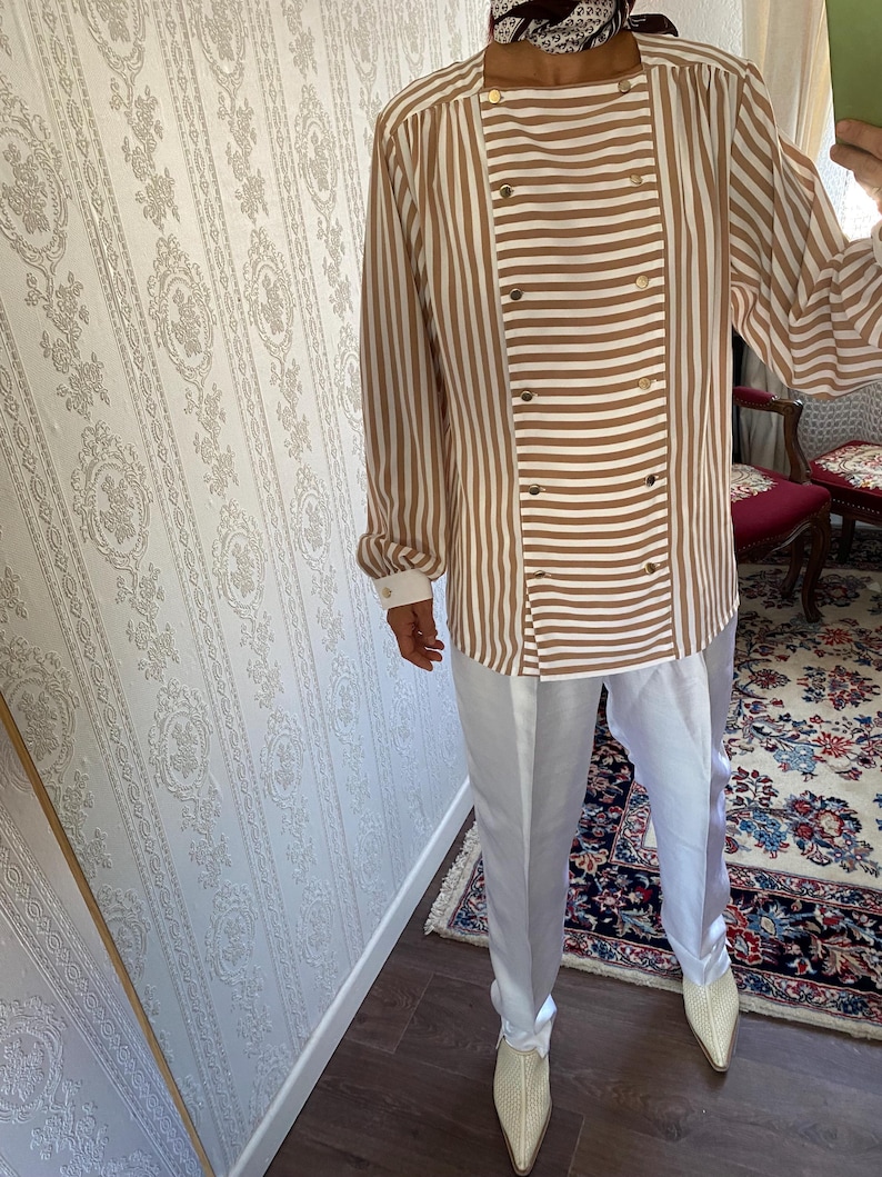 May include: A brown and white striped long-sleeve shirt with a double-breasted button closure. The shirt has a relaxed fit and is paired with white pants.