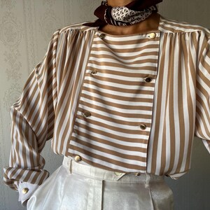May include: A brown and white striped blouse with a double-breasted closure and long sleeves. The blouse has a relaxed fit and a slightly oversized silhouette.