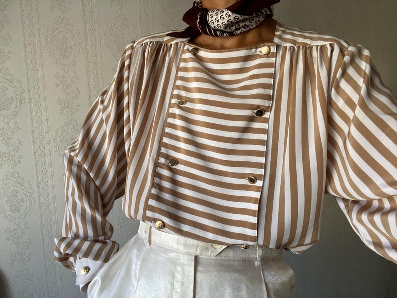 May include: A brown and white striped blouse with a double-breasted closure and long sleeves. The blouse has a relaxed fit and a slightly oversized silhouette.