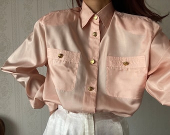 May include: A light pink satin button-down shirt with long sleeves and gold buttons. The shirt has two chest pockets with a flap.