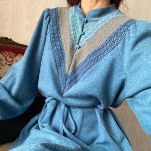 Peut inclure: Robe vintage bleu clair avec manches longues et taille nouée. Le corsage présente une encolure en V avec un panneau à motifs dans les tons de bleu et de beige. La robe a un col montant et trois boutons bleus.