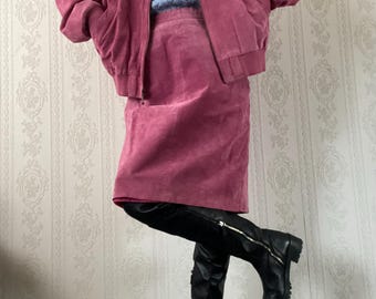 Puede incluir: Una chaqueta bomber de ante rosa y una falda a juego hasta la rodilla, sobre un suéter de cuello alto de punto trenzado azul claro. El conjunto se completa con botas negras hasta la rodilla con cremallera lateral y suela de goma gruesa.