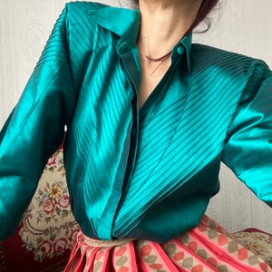 May include: A teal satin blouse with a pleated design and long sleeves. The blouse has a button-up front and a collar.