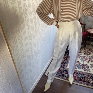 May include: A white striped blouse with brown stripes and gold buttons. The blouse has long sleeves and a relaxed fit. The model is wearing white pants and white shoes.