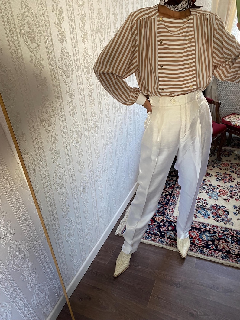 May include: A white striped blouse with brown stripes and gold buttons. The blouse has long sleeves and a relaxed fit. The model is wearing white pants and white shoes.