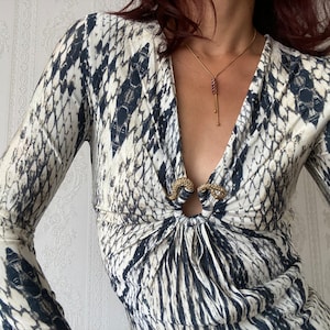May include: A long-sleeved, white and black patterned dress with a deep V-neckline. The dress has a gold-toned clasp at the neckline and a gold chain necklace with a dangling red gemstone pendant.
