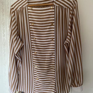 May include: A brown and white striped long-sleeved blouse with gold buttons. The blouse has a double-breasted closure and a collar.