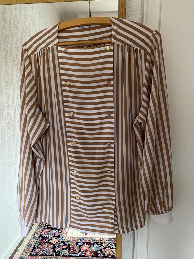 May include: A brown and white striped long-sleeved blouse with gold buttons. The blouse has a double-breasted closure and a collar.