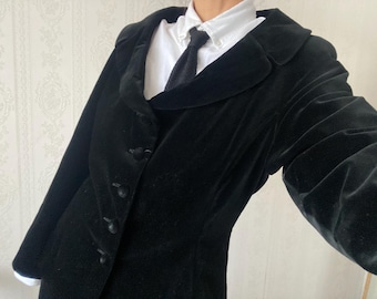 May include: A black velvet blazer with a notched lapel and button fastening. The blazer is worn over a white collared shirt and a black tie. The sleeves are long and the fabric appears soft.