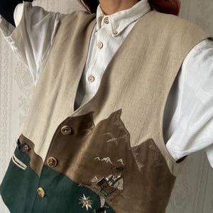 Puede incluir: Chaleco beige con una escena de montaña bordada en la parte delantera. El chaleco tiene un panel verde en el lado inferior izquierdo y botones dorados. El chaleco se lleva sobre una camisa blanca abotonada.