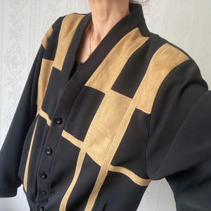 May include: Black cardigan with tan suede-like geometric design. The jacket has a V-neck, long sleeves, and a button-up front. The tan sections create a striking contrast against the black fabric, offering a bold, modern look. A stylish outerwear piece.