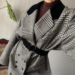 Puede incluir: Una chaqueta de cuadros de pata de gallo en blanco y negro con cuello de terciopelo negro y cinturón negro con hebilla dorada.