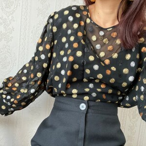 May include: A black sheer blouse with gold, silver, and copper polka dots. The blouse has long sleeves and a V-neckline. The model is wearing the blouse with black pants.