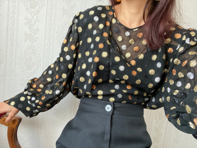May include: A black sheer blouse with gold, silver, and copper polka dots. The blouse has long sleeves and a V-neckline. The model is wearing the blouse with black pants.