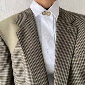 Puede incluir: Una blazer a cuadros marrón y beige con un parche beige en el hombro. La blazer se lleva sobre una camisa blanca con bordados florales y dos botones decorativos. La blazer tiene un corte clásico y entallado.
