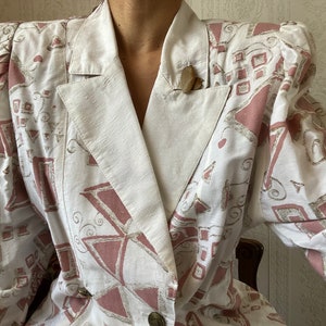 Op de afbeelding: Een vintage witte blazer met een geometrisch patroon in roze en grijstinten. De blazer heeft een revers, schoudervullingen en een sluiting met twee knopen. Een kleine houten speld zit in de zak.