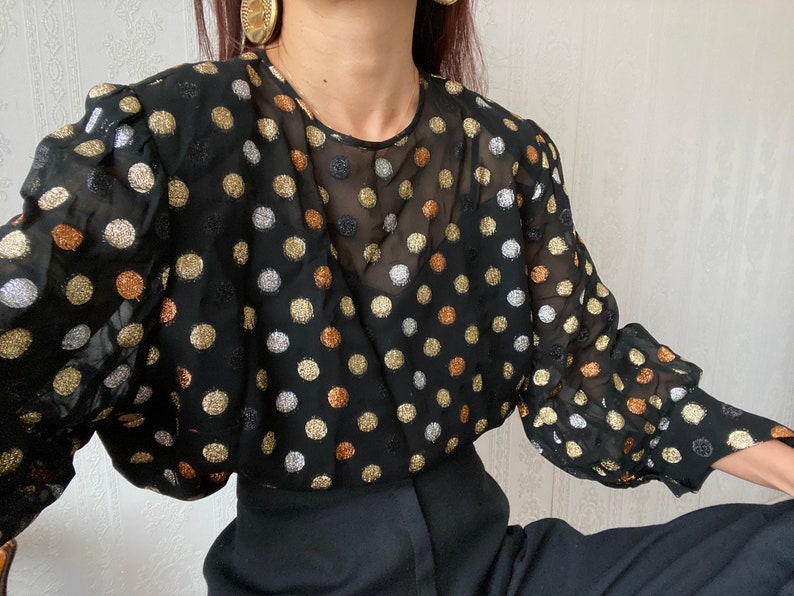 May include: A black sheer blouse with gold, silver, and copper polka dots. The blouse has long sleeves with a gathered detail at the shoulders.