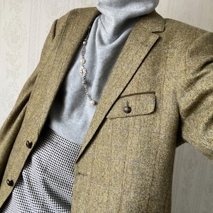 Può includere: Un blazer in tweed marrone con un bordo in pelle marrone sulla tasca. Il blazer è indossato sopra un maglione a collo alto grigio e una collana di perle bianche.