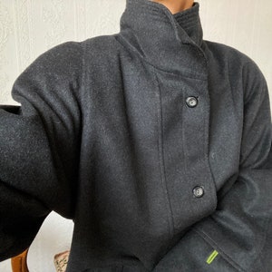 Puede incluir: Un abrigo de lana gris oscuro con cuello alto y cierre de botones. El abrigo tiene un corte holgado y mangas largas.