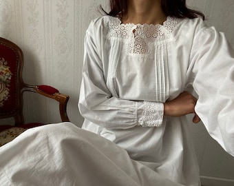 Può includere: Un camicione bianco in cotone con un delicato pizzo intorno al collo e ai polsini. Il camicione ha un corpetto drappeggiato e maniche lunghe.