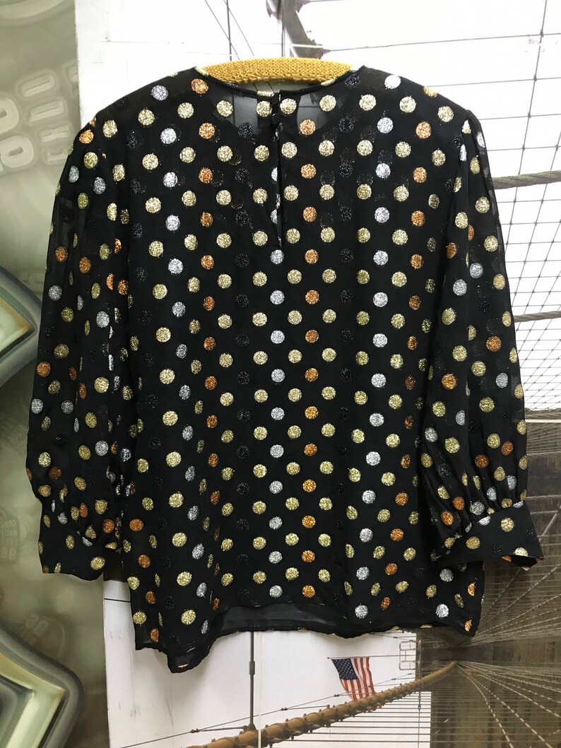 May include: A black sheer blouse with a round neckline and long sleeves. The blouse is covered in a polka dot pattern with gold, silver, and copper colored dots.